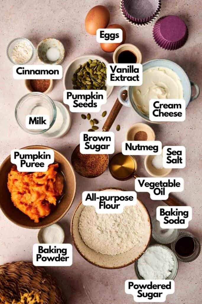 Ingredients for pumpkin muffins labeled on a table, including flour, pumpkin puree, baking soda, spices, and cream cheese.