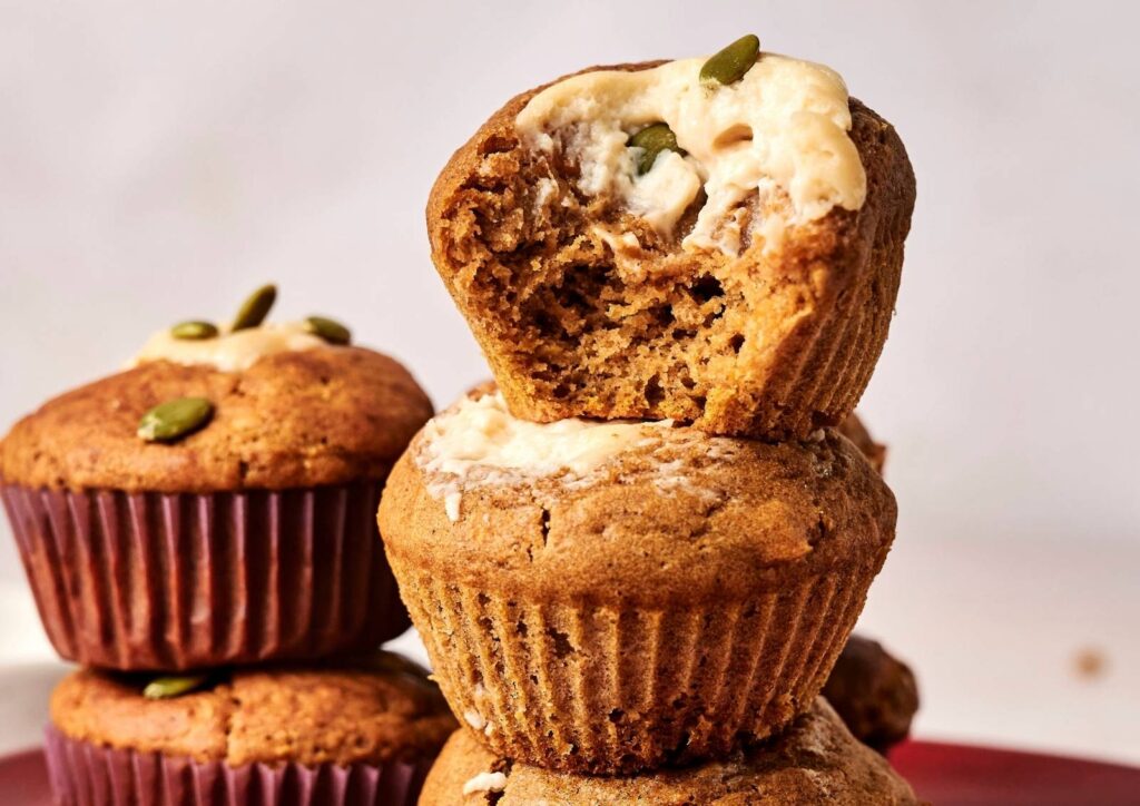 Stack of pumpkin cream cheese muffins with a bite taken out of the top one, showing the creamy filling and pumpkin seeds
