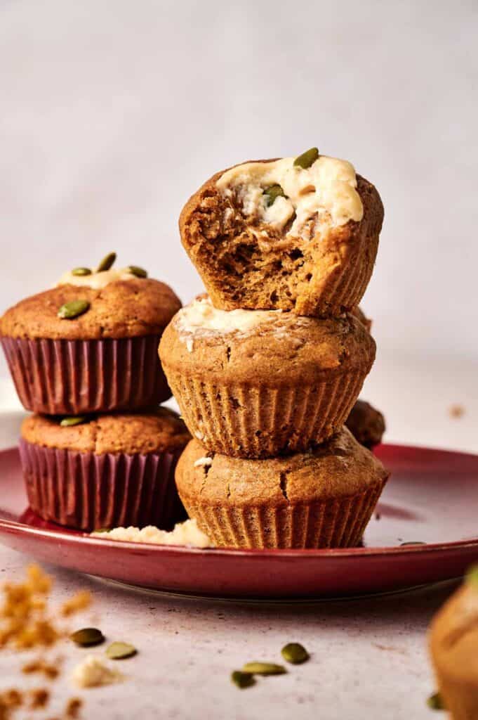 A stack of three muffins on a red plate, with the top muffin partially eaten. Pumpkin seeds are scattered around.
