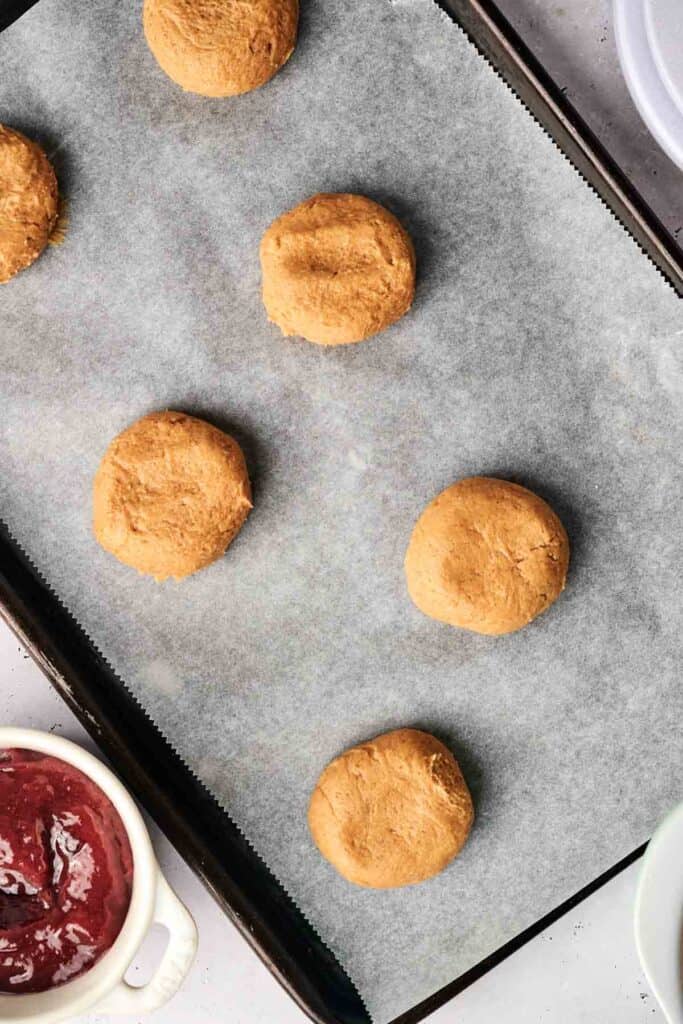 Six cookie dough balls on a baking sheet lined with parchment paper, reminiscent of Copycat Crumbl Peanut Butter and Jelly Cookies. A bowl with reddish jam-like contents is visible in the corner.