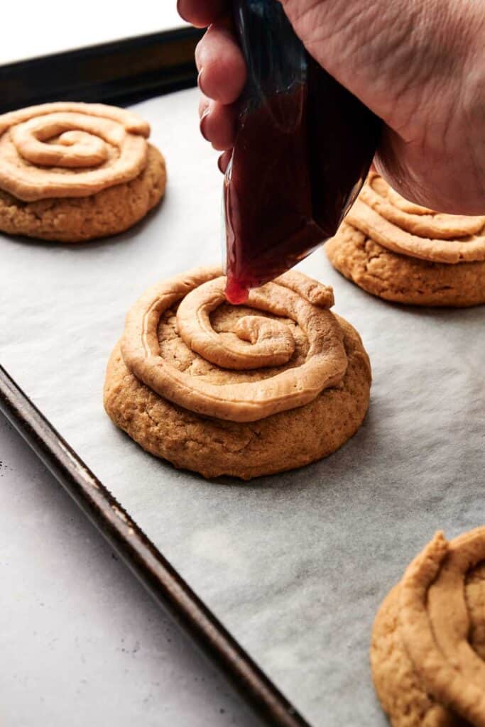 A hand pipes filling onto large round cookies on a parchment-lined baking sheet, creating delectable Copycat Crumbl Peanut Butter and Jelly Cookies.