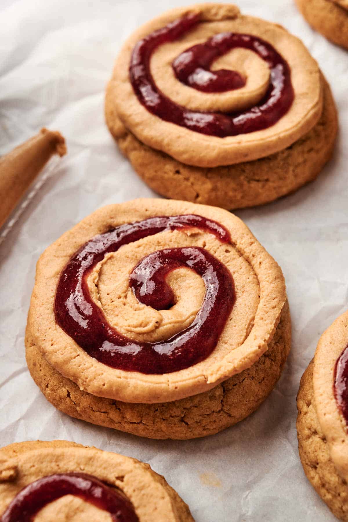 Close-up of freshly baked Copycat Crumbl Peanut Butter and Jelly Cookies with a swirl of red jam on top, placed on parchment paper.
