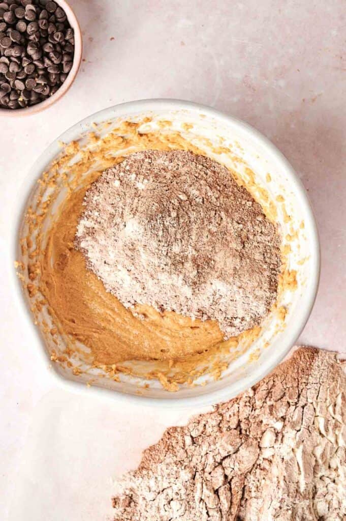 A bowl of cookie dough with dry ingredients sits ready, channeling the essence of Copycat Crumbl Chocolate Cake Cookies. Nearby, a small bowl brims with luscious chocolate chips, promising a treat to remember.