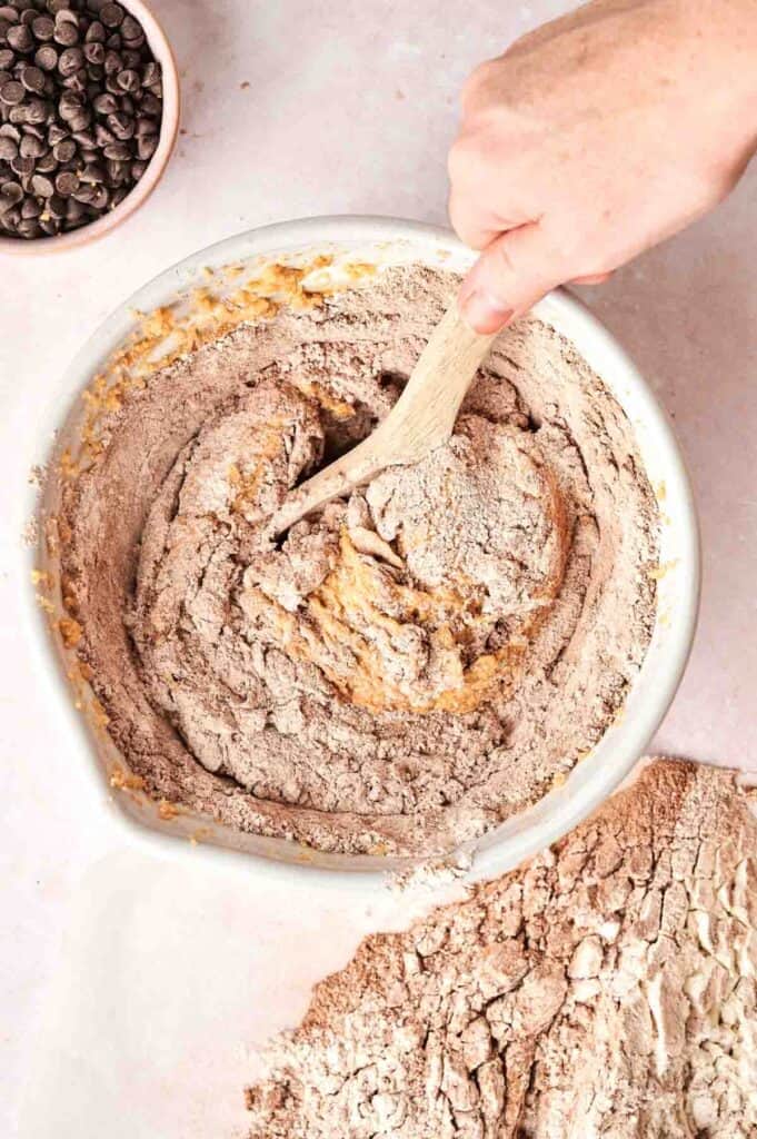 A hand mixing chocolate batter in a bowl with a wooden spoon, surrounded by spilled flour, captures the essence of baking copycat Crumbl Chocolate Cake Cookies. A bowl of chocolate chips sits invitingly in the corner.