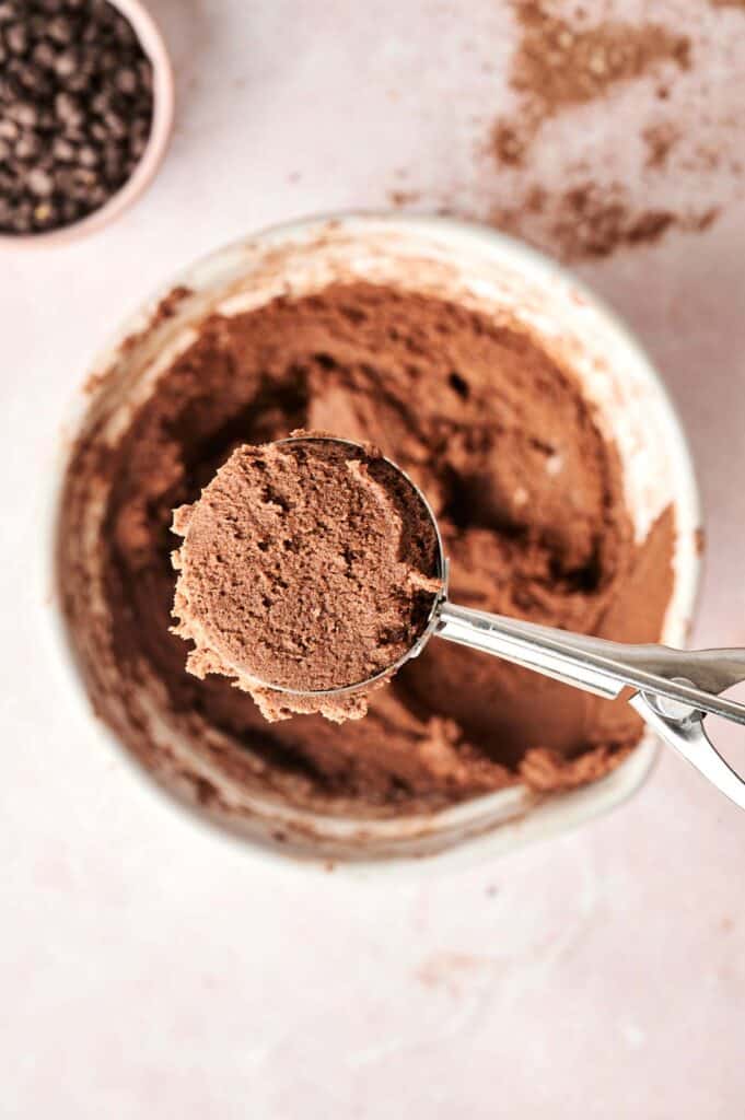 A scoop of copycat Crumbl chocolate cake cookie dough rests in a metal scooper poised above the mixing bowl, while chocolate chips sit temptingly in a small bowl in the background.