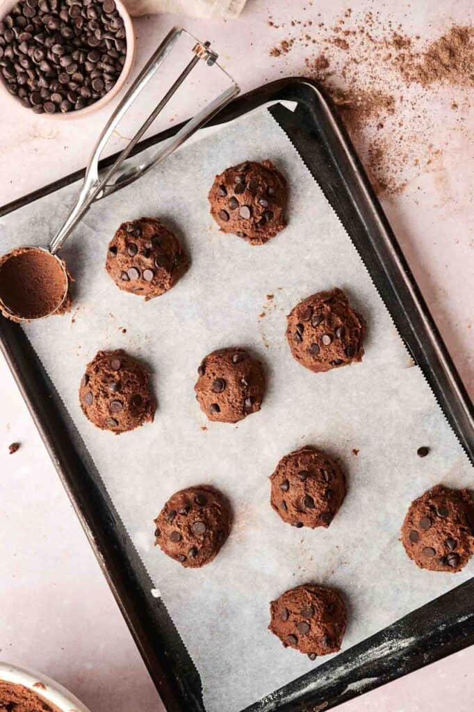 Unbaked Copycat Crumbl Chocolate Cake Cookies rest enticingly on a parchment-lined baking tray, accompanied by a trusty cookie scoop and a bowl of rich chocolate chips.
