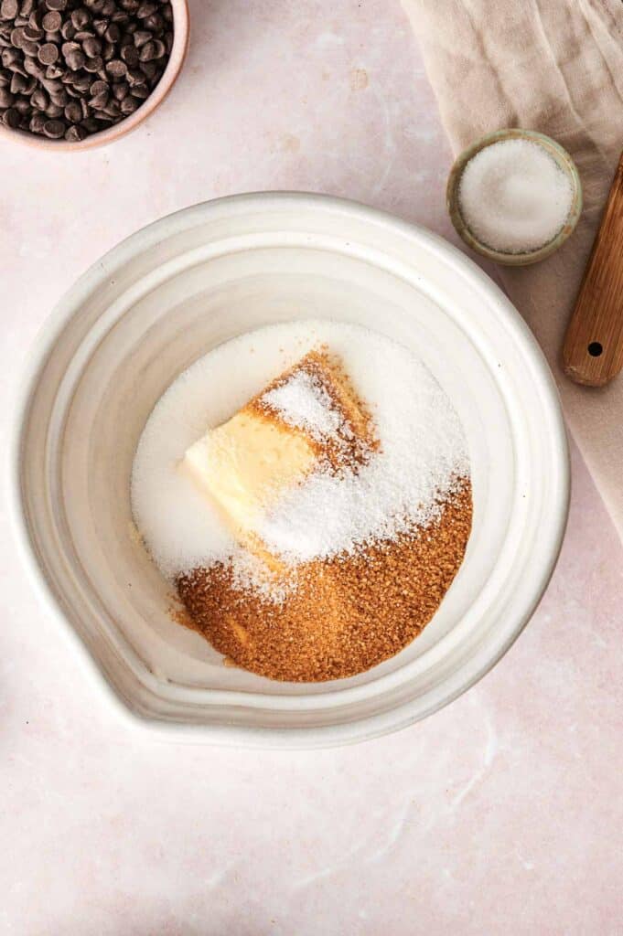 A bowl containing sugar, brown sugar, and butter sits invitingly on a light-colored surface, surrounded by chocolate chips and a small bowl of salt. Perfect for recreating those irresistible Copycat Crumbl Chocolate Cake Cookies at home.