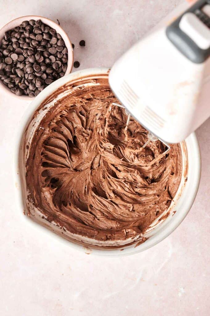 A bowl of copycat Crumbl chocolate cake batter is being mixed with an electric mixer, promising a delicious batch of cookies. A small bowl of chocolate chips is nearby, ready to be folded into the mix for that extra touch of indulgence.