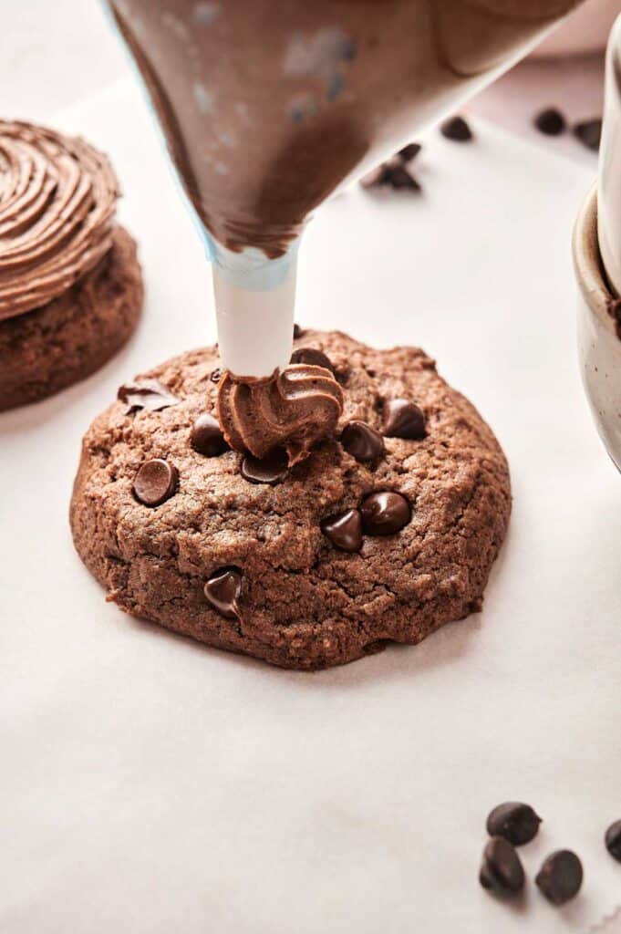 A chocolate cookie, reminiscent of copycat Crumbl Chocolate Cake Cookies, is elegantly decorated with a swirl of rich chocolate frosting using a piping bag. Chocolate chips are artfully scattered around on the white parchment paper.