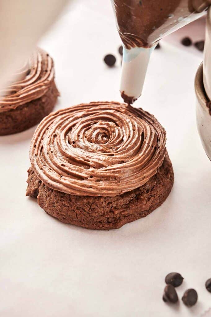 A piping bag expertly swirls chocolate frosting onto a chocolate cookie, reminiscent of Copycat Crumbl Chocolate Cake Cookies. Nearby, coffee beans are artfully scattered, adding an aromatic touch.