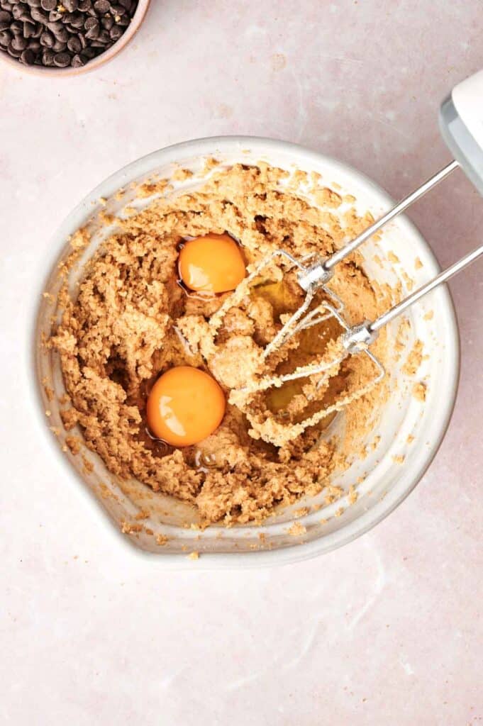 A mixing bowl with brown dough and two cracked eggs, accompanied by an electric mixer, sits ready for action. Nearby, a small bowl of chocolate chips waits on a light surface—perfect for crafting copycat Crumbl Chocolate Cake Cookies.