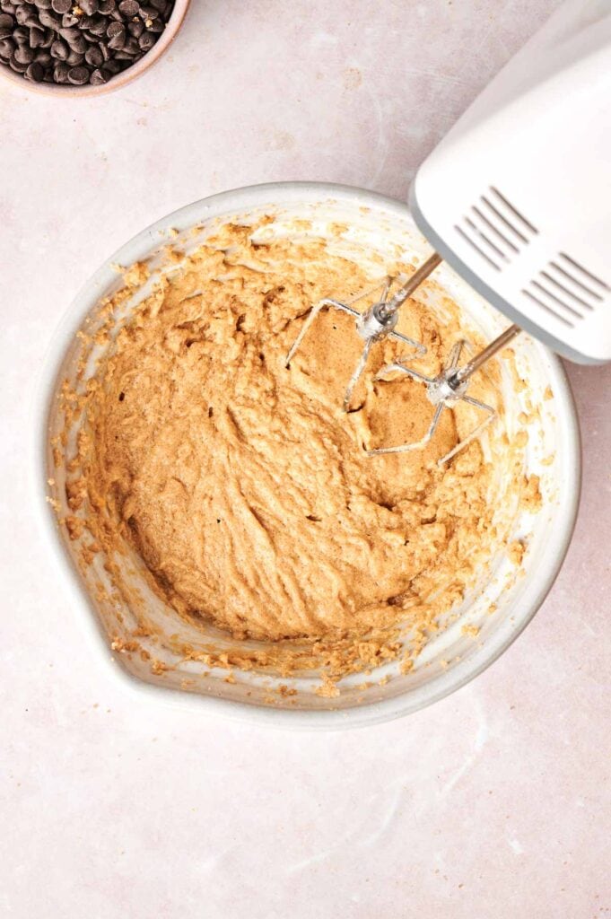 A mixing bowl holds a light brown batter with an electric hand mixer poised for action, ready to create copycat Crumbl chocolate cake cookies. Nearby, a small bowl brims with chocolate chips, waiting to lend their sweetness.
