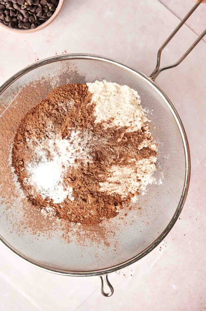 A sifter filled with a mix of cocoa powder, flour, and baking soda sits on a light-colored surface, alongside a small bowl of chocolate chips—perfect for crafting Copycat Crumbl Chocolate Cake Cookies.
