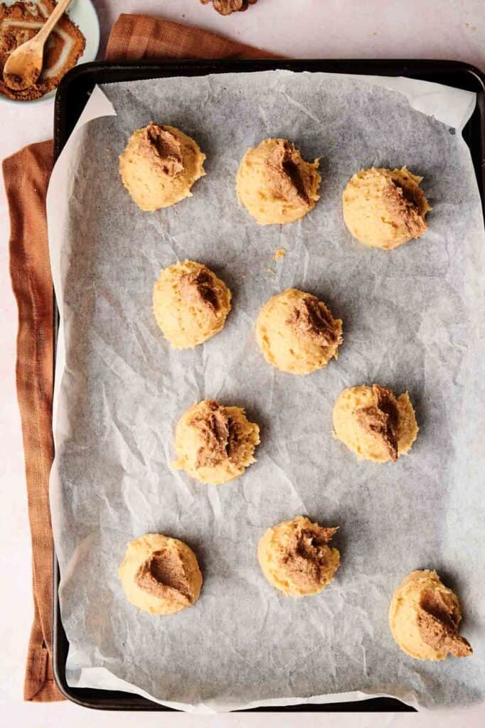 A baking tray lined with parchment paper holds nine unbaked Copycat Crumbl Cinnamon Swirl Cookies, each adorned with a spoonful of brown sugar topping. A cozy brown cloth rests beneath the tray, adding a warm touch to this irresistible recipe.
