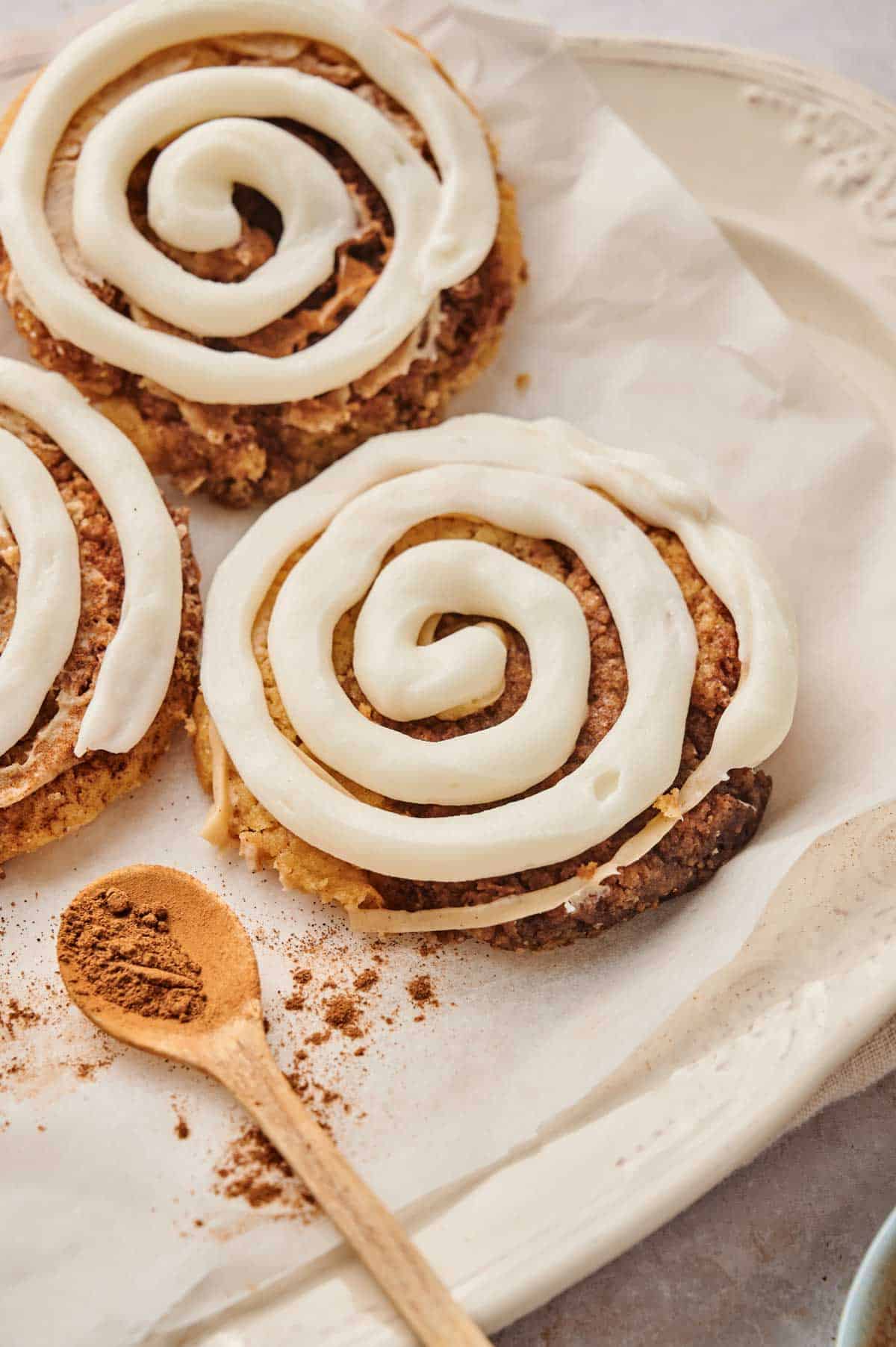 Three cinnamon rolls topped with white icing sit on a plate, reminiscent of a Copycat Crumbl Cinnamon Swirl Cookies recipe, with a wooden spoon holding ground cinnamon beside them.