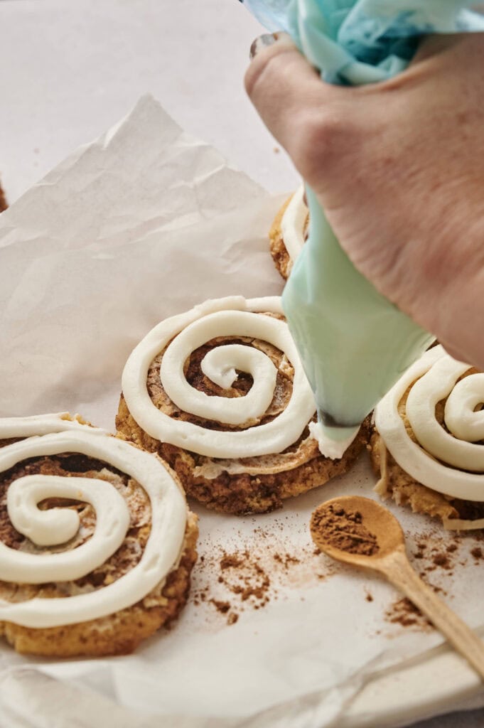 A hand uses a piping bag to add swirls of white icing on Copycat Crumbl Cinnamon Swirl Cookies placed on parchment paper, with a wooden spoon and cinnamon nearby.