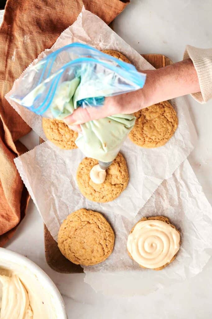 A person is piping green icing onto cookies with a clear plastic bag, on a parchment-lined surface with more cookies nearby.