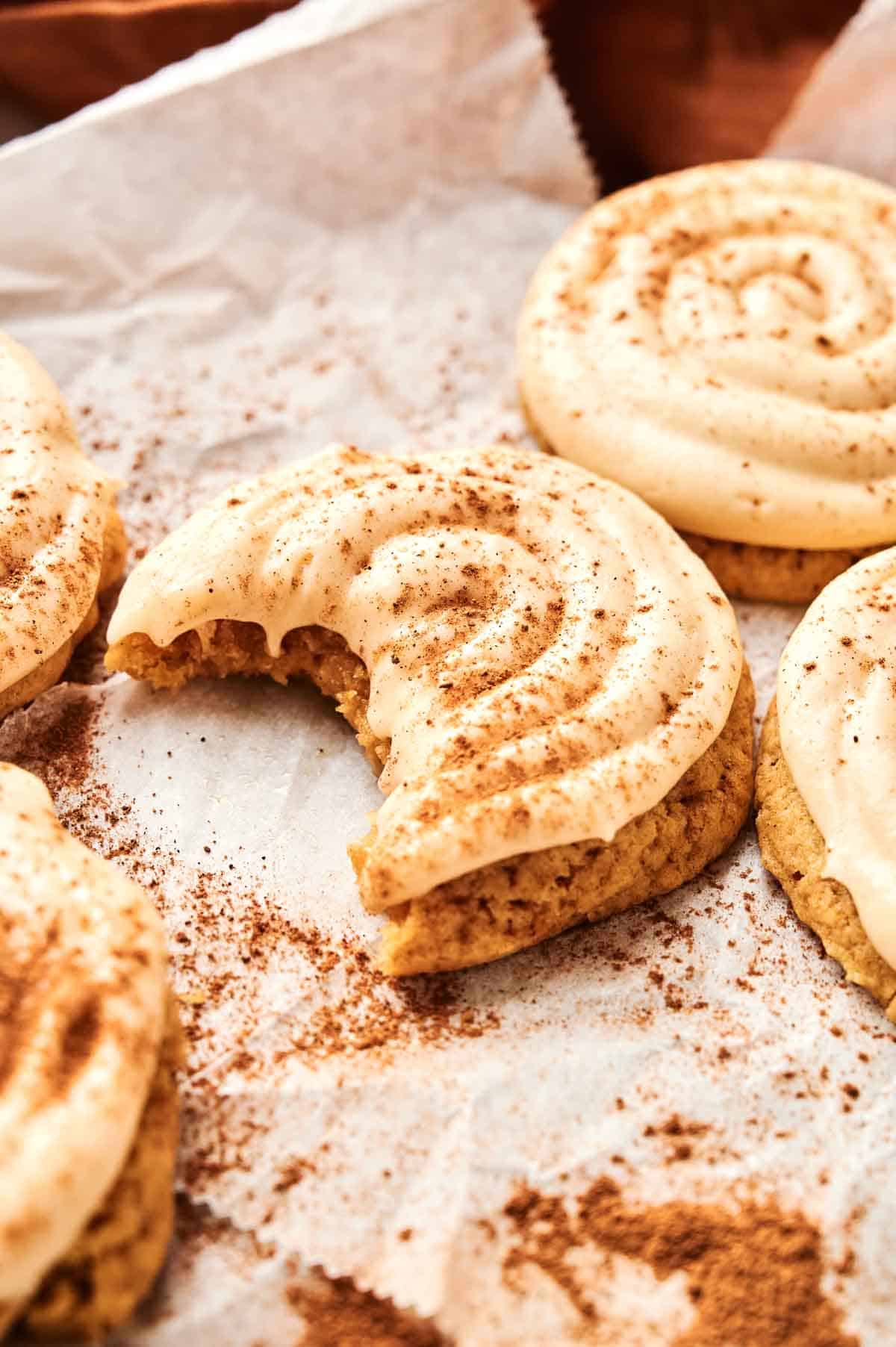 Freshly baked cookies on parchment paper, topped with white frosting and a sprinkle of cinnamon. One cookie has a bite taken out of it.