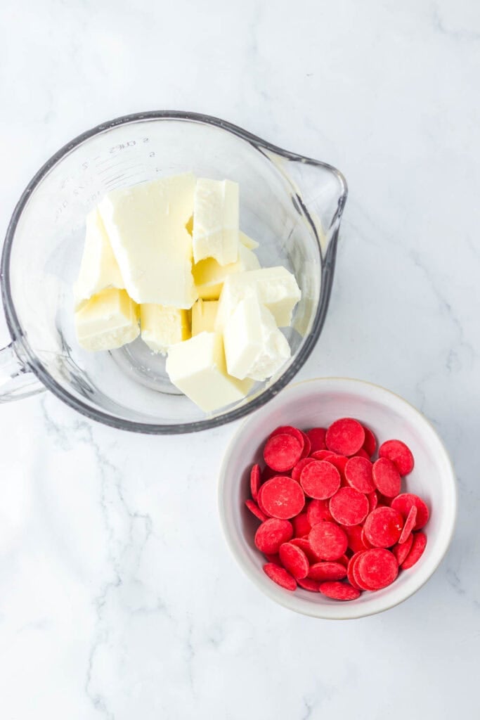 A glass bowl with white chocolate chunks and a small white bowl with red chocolate wafers, resembling Spooky Halloween Eyeballs, rest on a marble countertop.