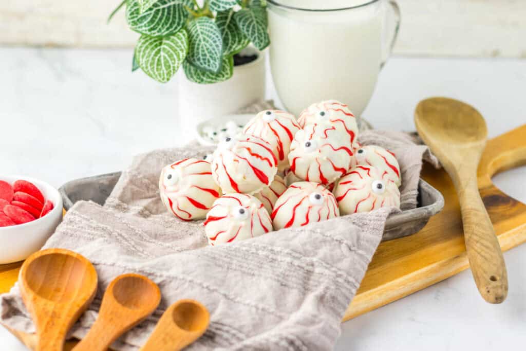 A tray of white chocolate truffles decorated to resemble spooky Halloween eyeballs with red lines and candy eyes, placed on a cloth next to wooden spoons, a glass of milk, and a potted plant in the background.