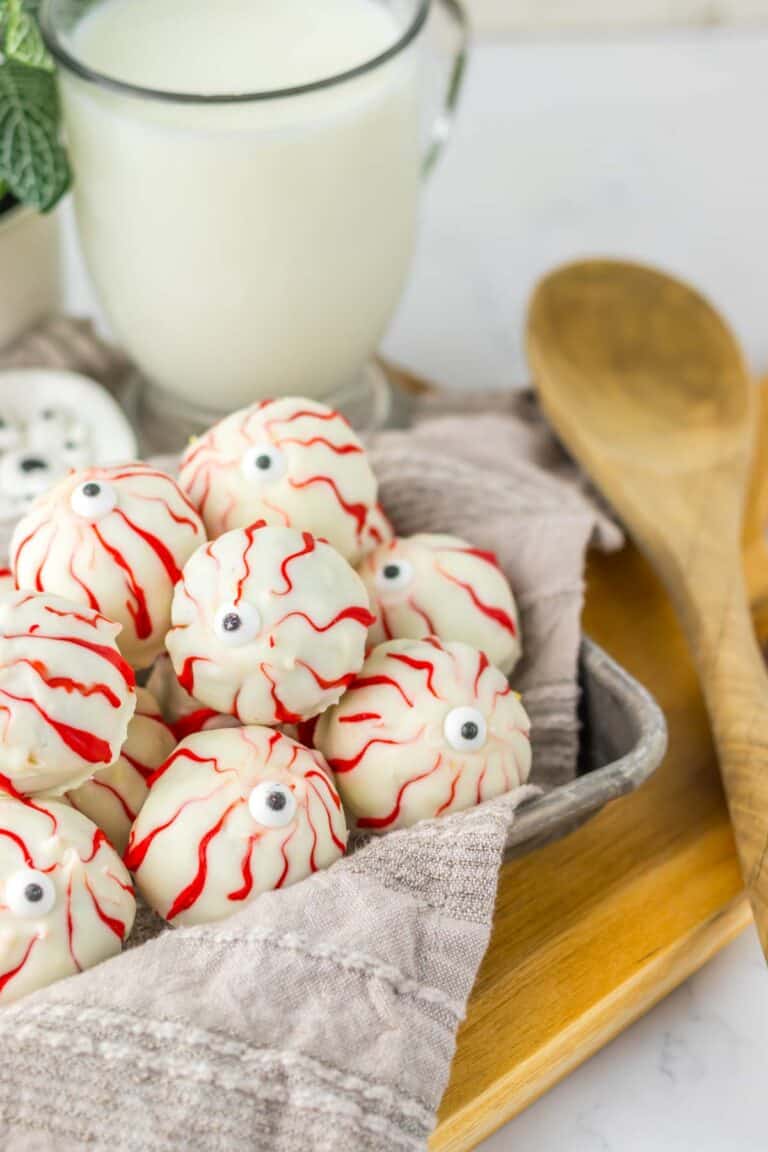 A tray of white chocolate truffles decorated to resemble spooky Halloween eyeballs, with red icing and candy eyes, accompanied by a glass of milk and wooden spoons.
