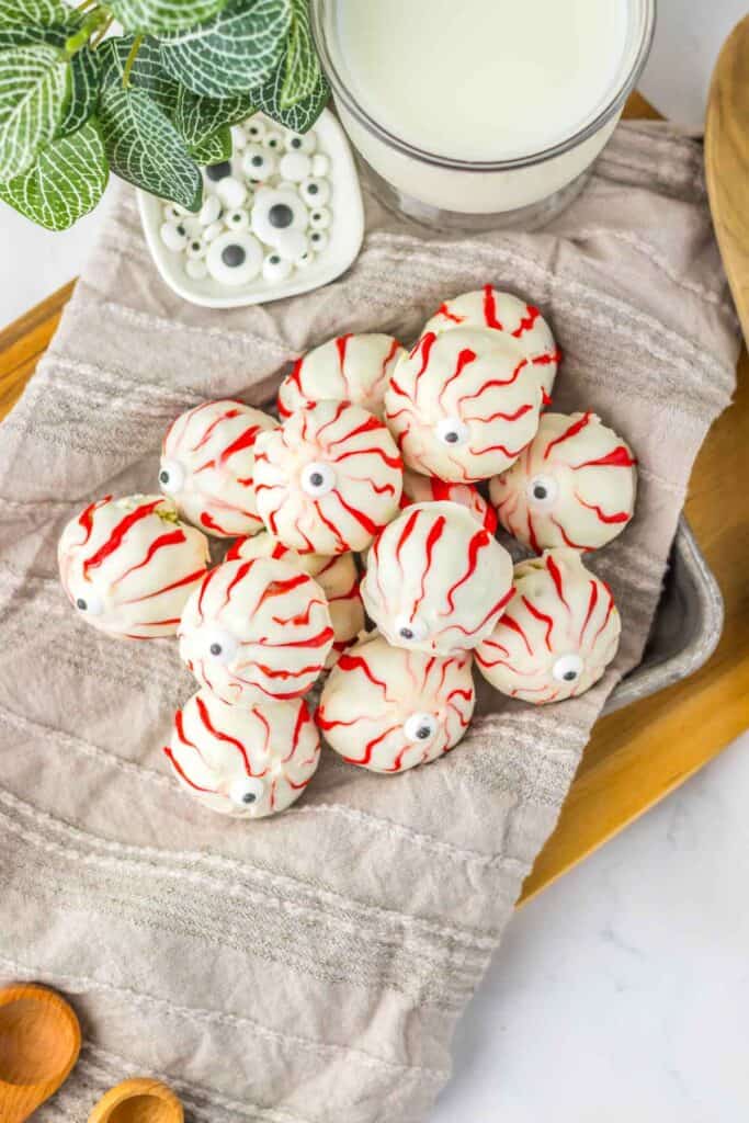 A tray holding several spooky Halloween eyeballs—white confections with red veining and a central black dot, accompanied by a plant and a glass of milk in the background.