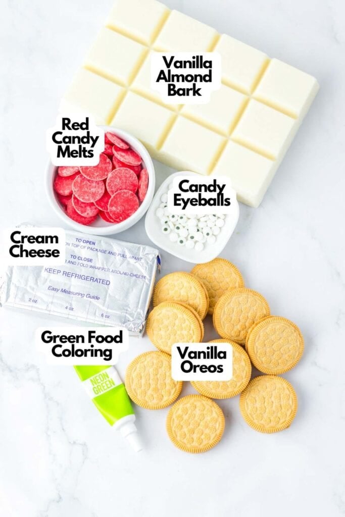 Ingredients for a dessert include vanilla almond bark, red candy melts, spooky Halloween eyeballs candy, cream cheese, green food coloring, and vanilla Oreos on a white background.