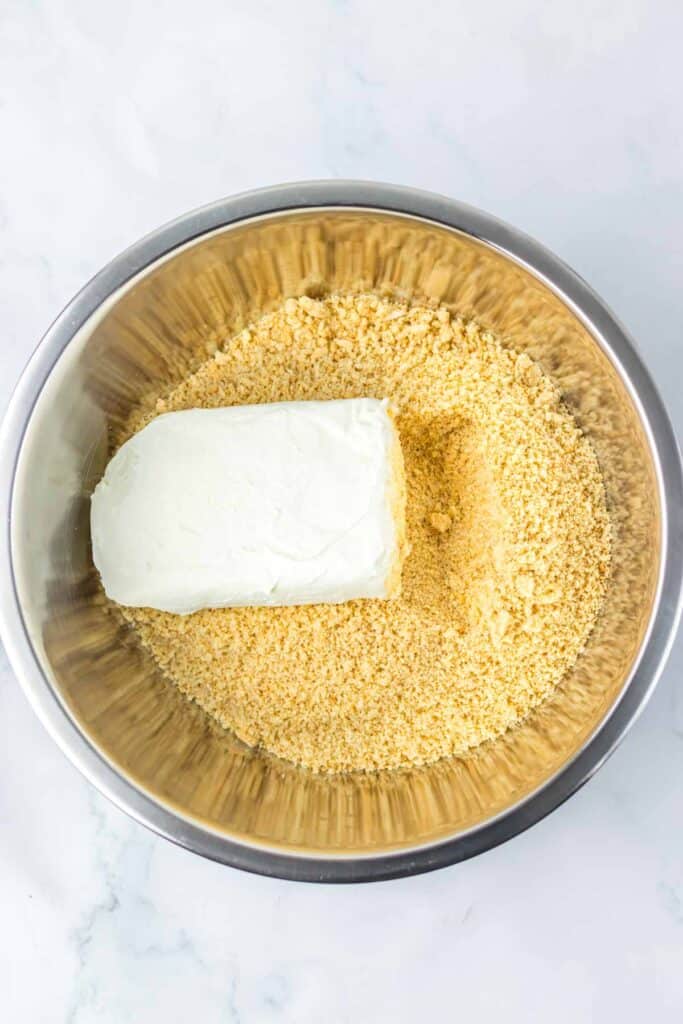 A block of cream cheese sits on a bed of crushed graham crackers in a stainless steel mixing bowl, ready to be transformed into spooky Halloween eyeballs.