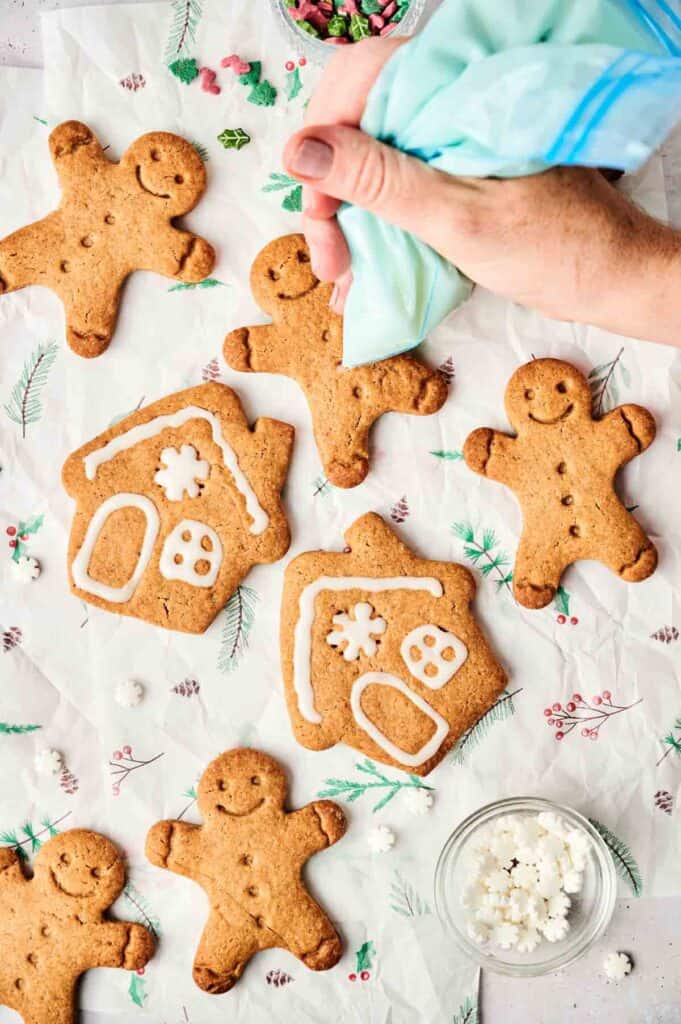 Hand decorating gingerbread cookies with white icing on a festive tablecloth. Gingerbread men and houses are visible, along with small bowls of decorations.