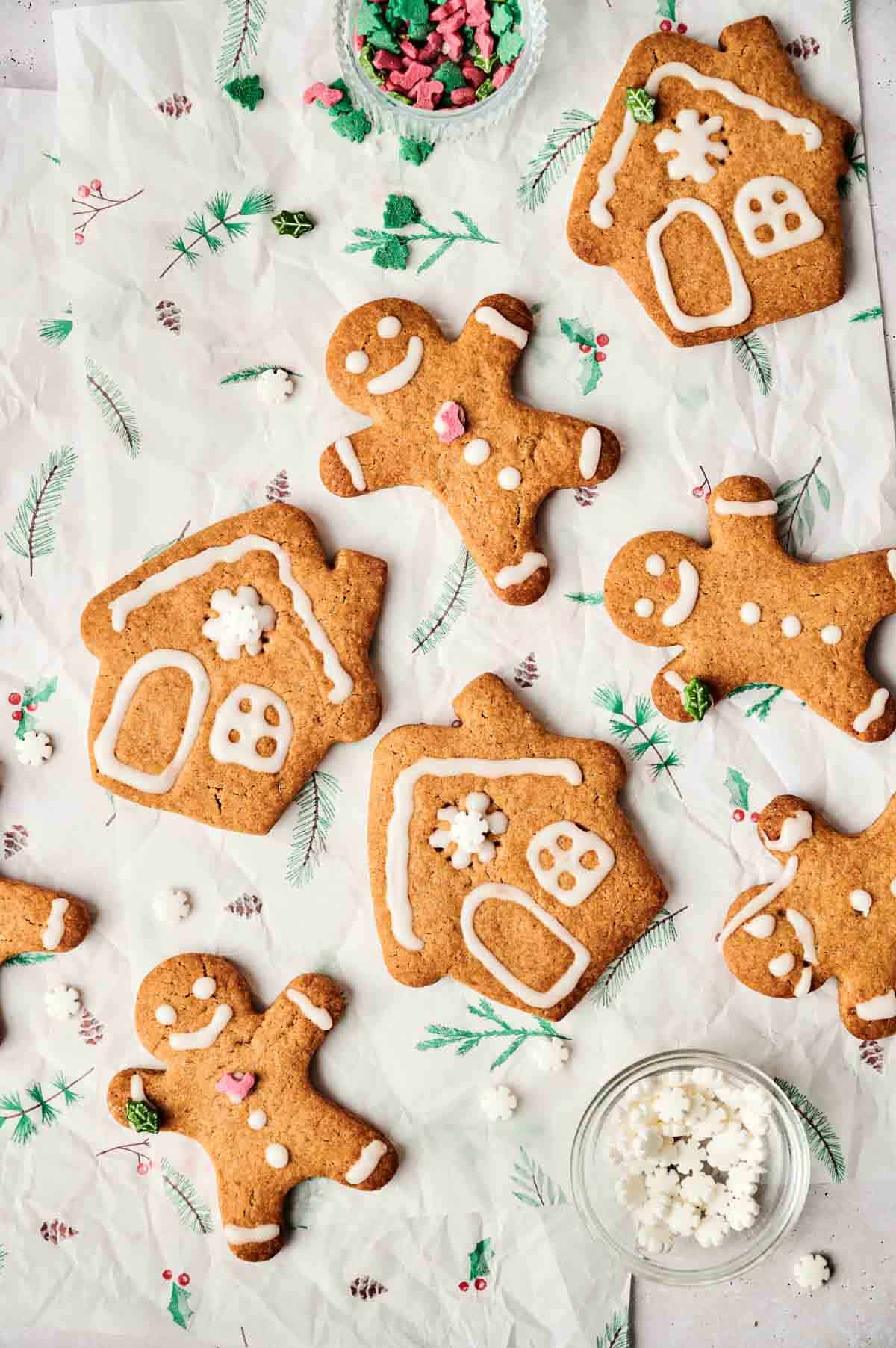 Assorted gingerbread cookies shaped like houses and people are decorated with icing on a festive paper backdrop. Small bowls of star-shaped sprinkles and decorations are nearby.