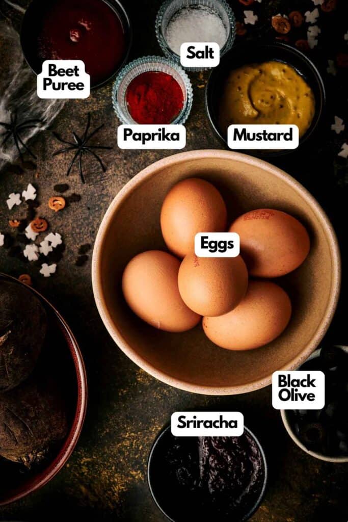 Ingredients arranged on a table for Halloween deviled eggs include eggs, beet puree, salt, paprika, mustard, black olives, and sriracha. Spooky decorations add to the festive atmosphere.