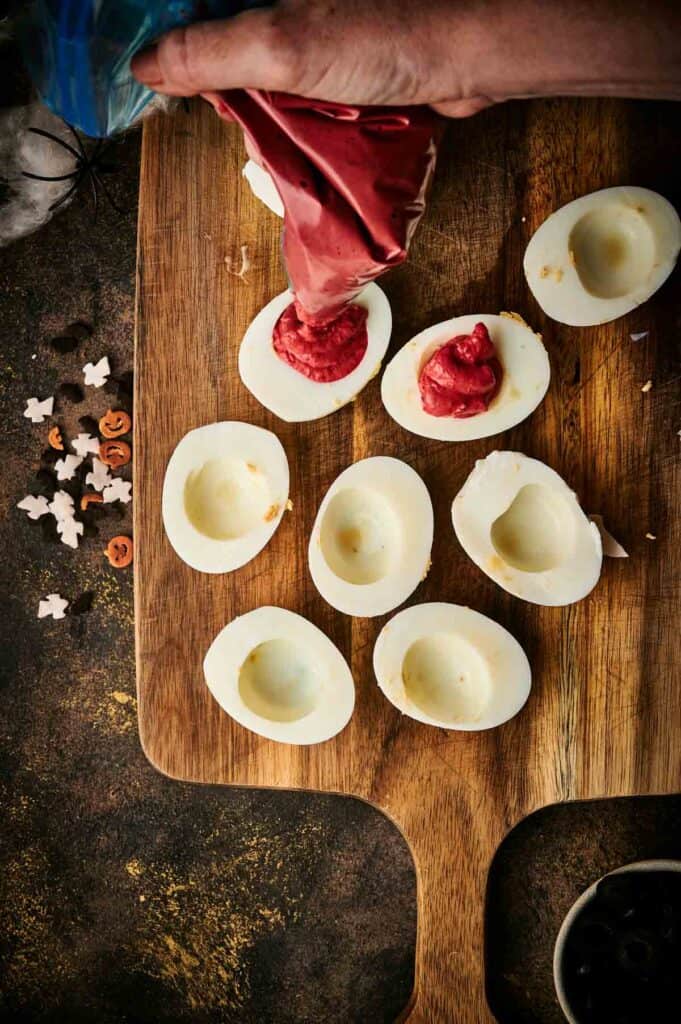 A hand expertly fills hollowed hard-boiled egg halves with a red filling using a piping bag on a wooden board, crafting spooky Halloween deviled eggs. Eerie decorations are scattered nearby, perfecting the festive atmosphere.