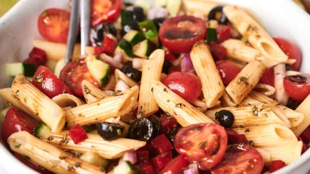 A bowl of pasta salad with penne, cherry tomatoes, black olives, cucumbers, and diced vegetables, garnished with herbs.