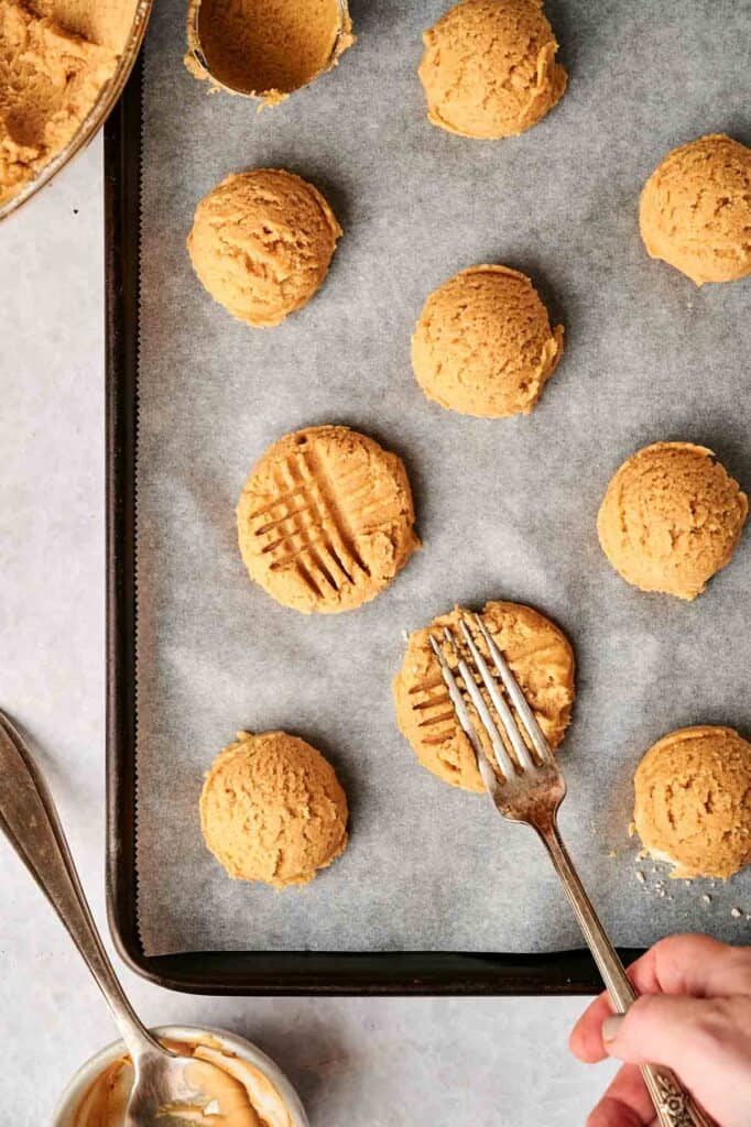 Cookie dough balls on a baking sheet, some with fork marks. A hand uses a fork to press patterns onto the dough.