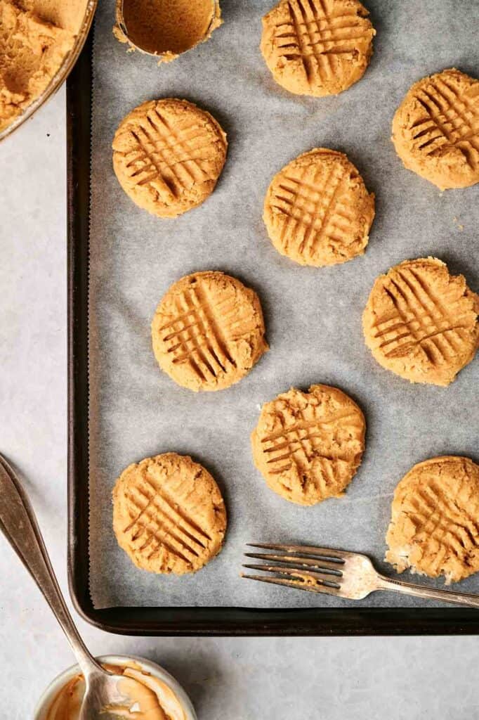 Baking sheet with unbaked peanut butter cookies, each with a crisscross fork pattern on top. A fork and a small bowl nearby.