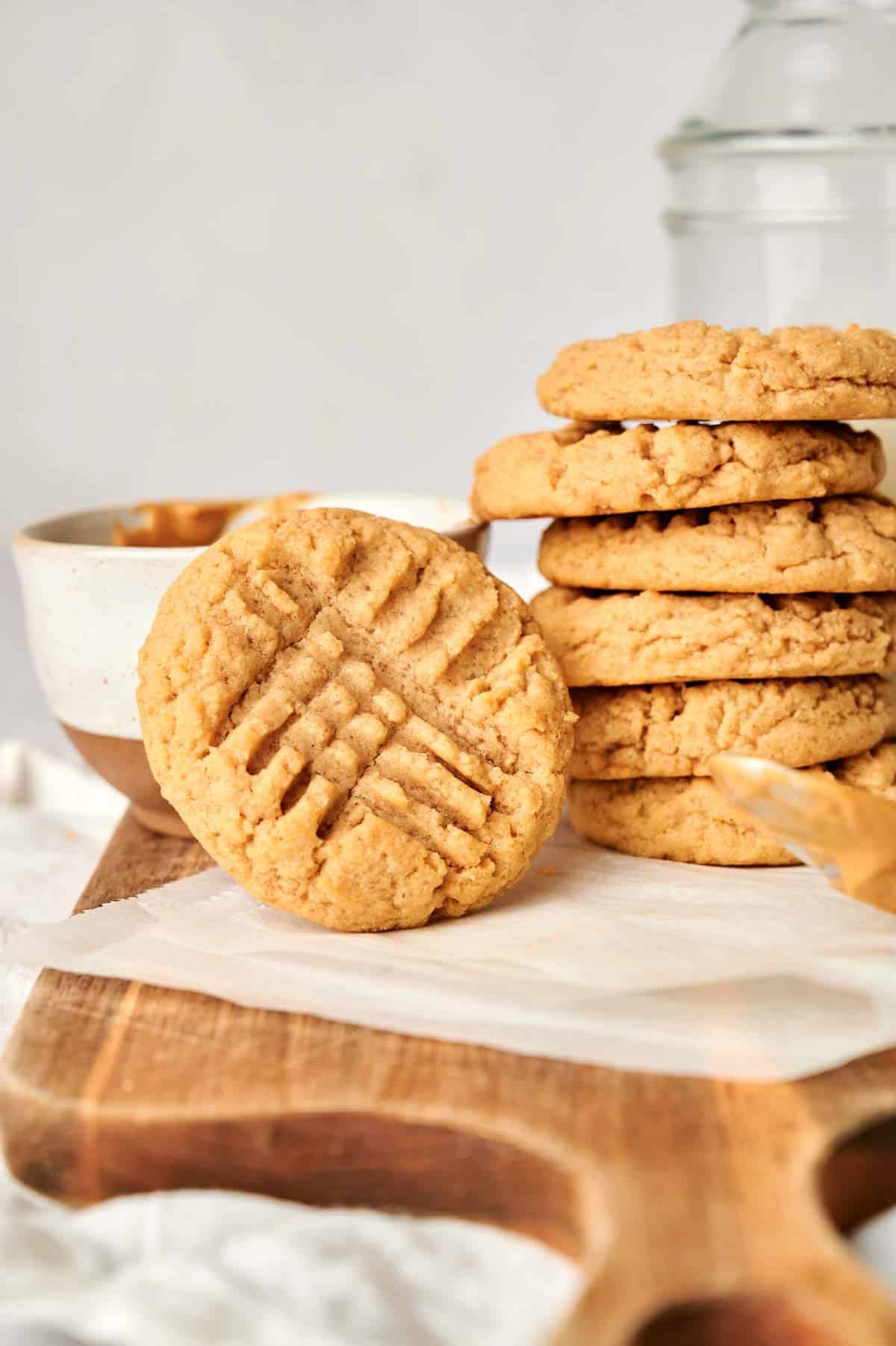 A stack of peanut butter cookies on a wooden board, with one cookie leaning against it. A bowl and knife with peanut butter are in the background.