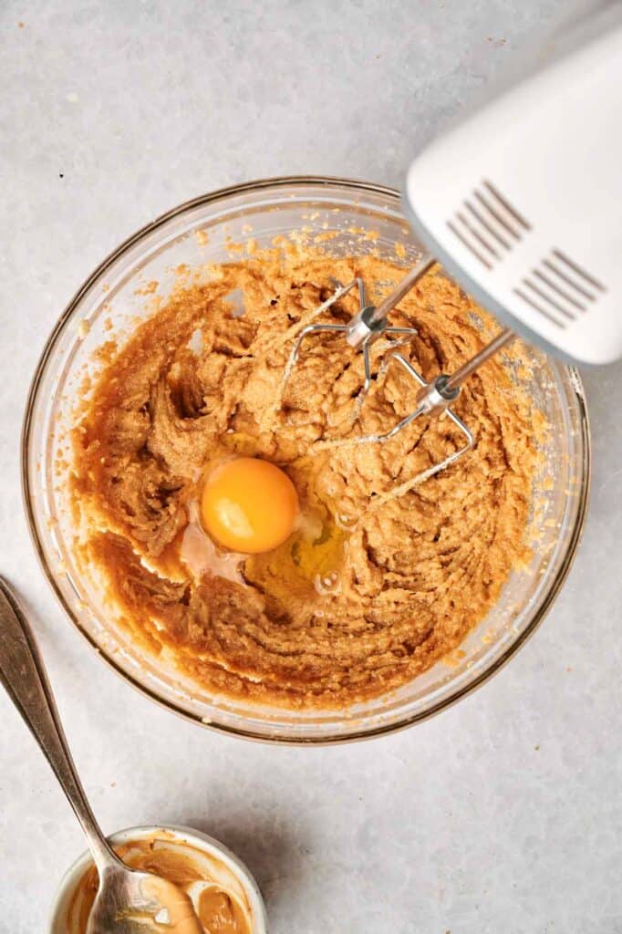 A bowl with brown dough mixture and a single egg in the center. An electric mixer and a spoon are nearby.