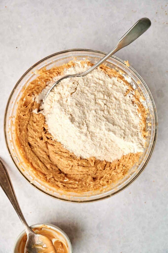 A glass bowl with cookie dough and a pile of flour on top, with a spoon in the mixture, and a small bowl of peanut butter nearby.