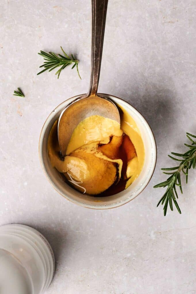 A bowl with a creamy brown sauce, perfect for pairing with turkey breast, and a spoon, garnished with rosemary sprigs, sits on a light stone surface.