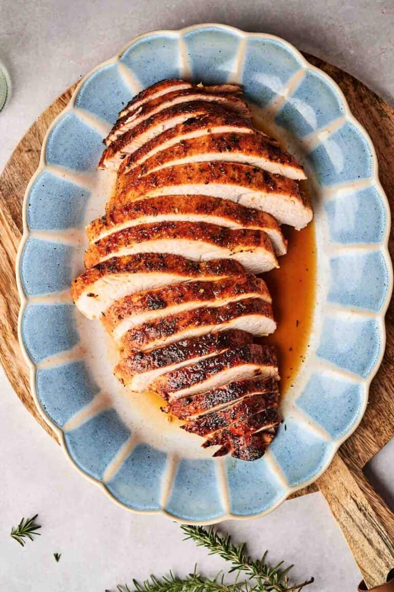 Sliced roasted turkey breast on a blue oval platter with visible seasoning and juice, set on a wooden surface. Nearby, sprigs of rosemary add a touch of elegance.