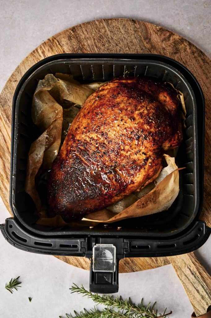 Cooked seasoned chicken breast in an air fryer basket on a wooden board, garnished with sprigs of rosemary.