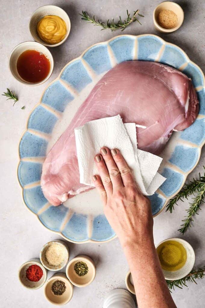 A hand pats raw pork tenderloin dry with a paper towel on a blue-rimmed platter, surrounded by small bowls of spices, herbs, and mustard. Nearby, a succulent turkey breast awaits similar preparation.