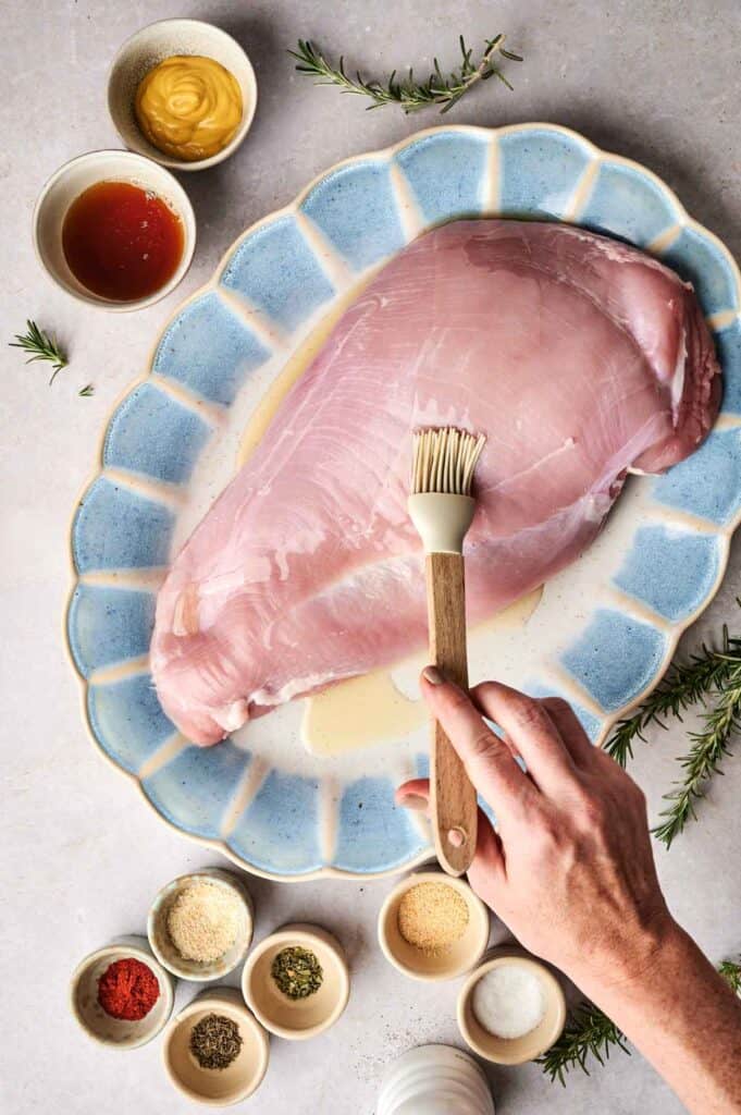 A raw turkey breast sits on a blue platter, being brushed with marinade. Small bowls of spices, herbs, mustard, and sauce surround it, while rosemary sprigs rest beside the platter.