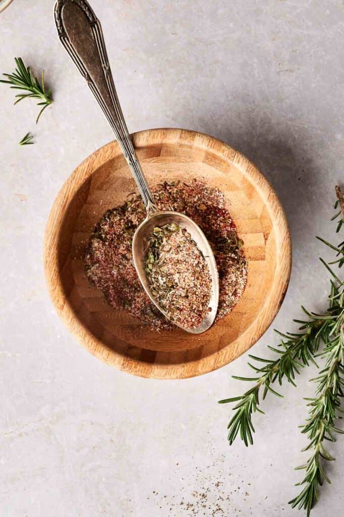 A wooden bowl filled with mixed herbs and spices sits next to a spoon on a light surface, perfect for seasoning turkey breast. Nearby, sprigs of rosemary add a fragrant touch.