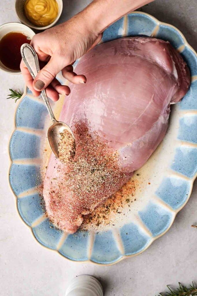 A person sprinkles seasonings on a large turkey breast atop a blue-rimmed platter. Nearby, bowls of mustard and a rich brown sauce await, adding aromatic anticipation to this flavorful preparation.