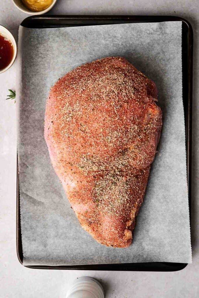 Raw seasoned turkey breast rests on a baking sheet lined with parchment paper, accompanied by small bowls of mustard and sauce nearby.