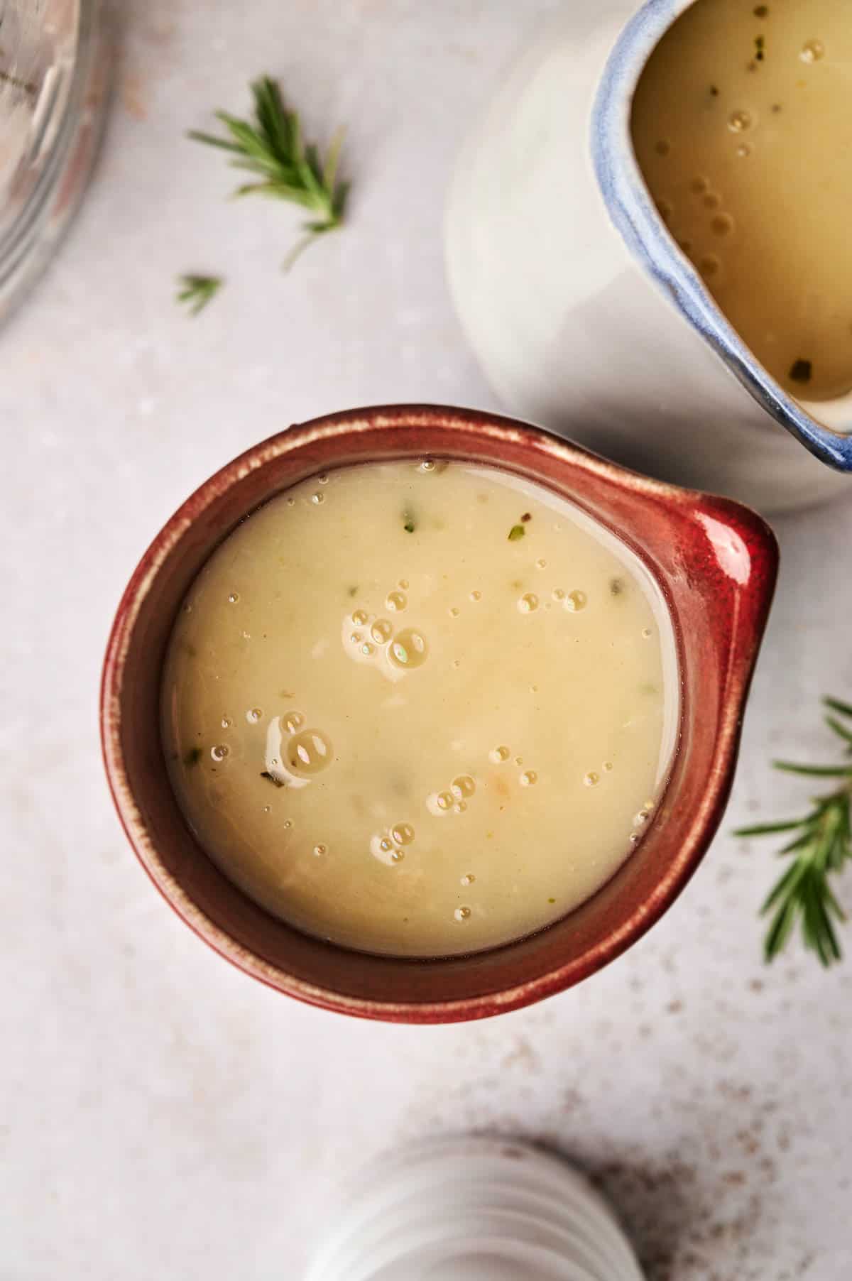 A red jug brimming with creamy turkey gravy is surrounded by fresh green herbs on a light surface.