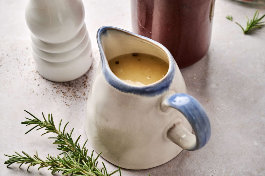 A ceramic gravy boat filled with light brown turkey gravy sits on a countertop, accompanied by a sprig of rosemary and a white pepper grinder nearby.