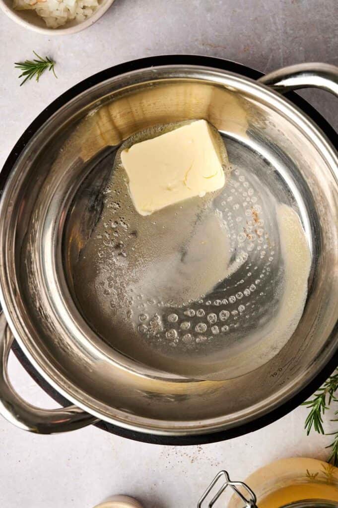 A pat of butter melting in a stainless steel pot on a light-colored surface, preparing the base for turkey gravy, with small rosemary sprigs nearby.