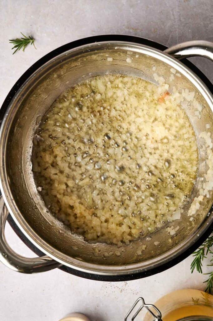 Chopped onions saut&eacute;ing in a pot with bubbling oil, setting the stage for a savory turkey gravy. A fresh rosemary sprig is visible in the background, promising aromatic depth to the sauce.